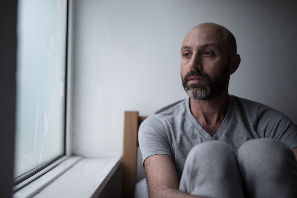 A bald man with anxiety sits on a bed, gazing thoughtfully out the window